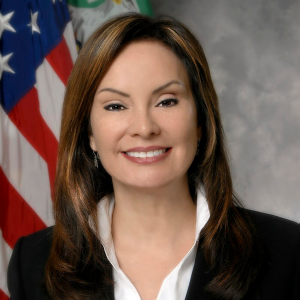 Headshot of Rosie Rios in black blazer with white shirt in front of American flag posted to her right. 