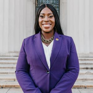 Photo of Vanessa L. Gibson, standing with hands in front of her, wearing a purple blazer. 